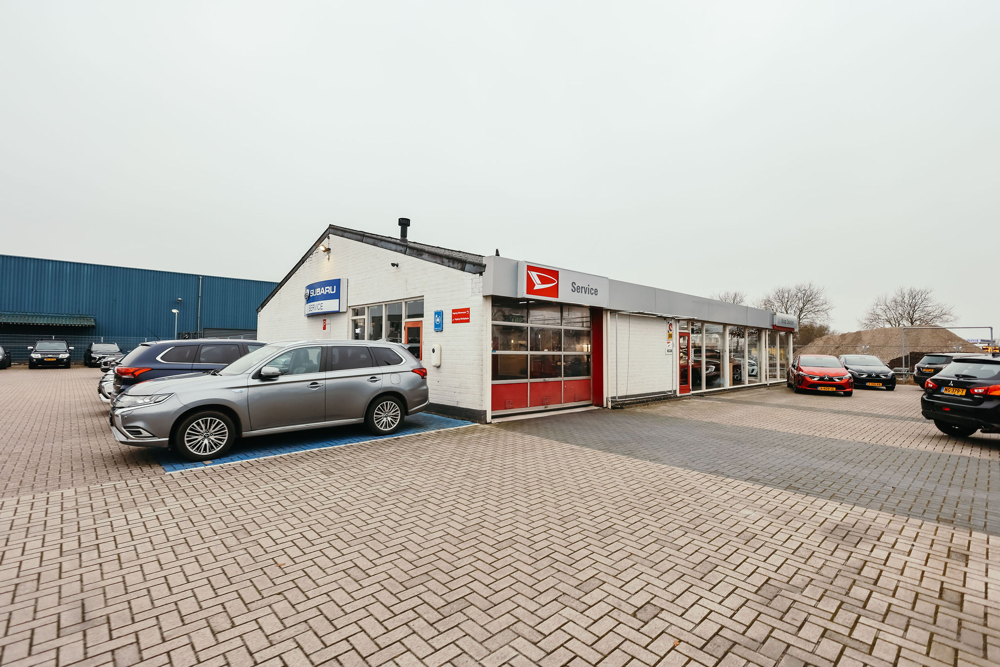 Autobedrijf Doorten in Hoogeveen met een Subaru Service-bord en Daihatsu Service-garage. Op de parkeerplaats staan meerdere auto's, waaronder een zilveren SUV, een rode hatchback en enkele zwarte auto's.