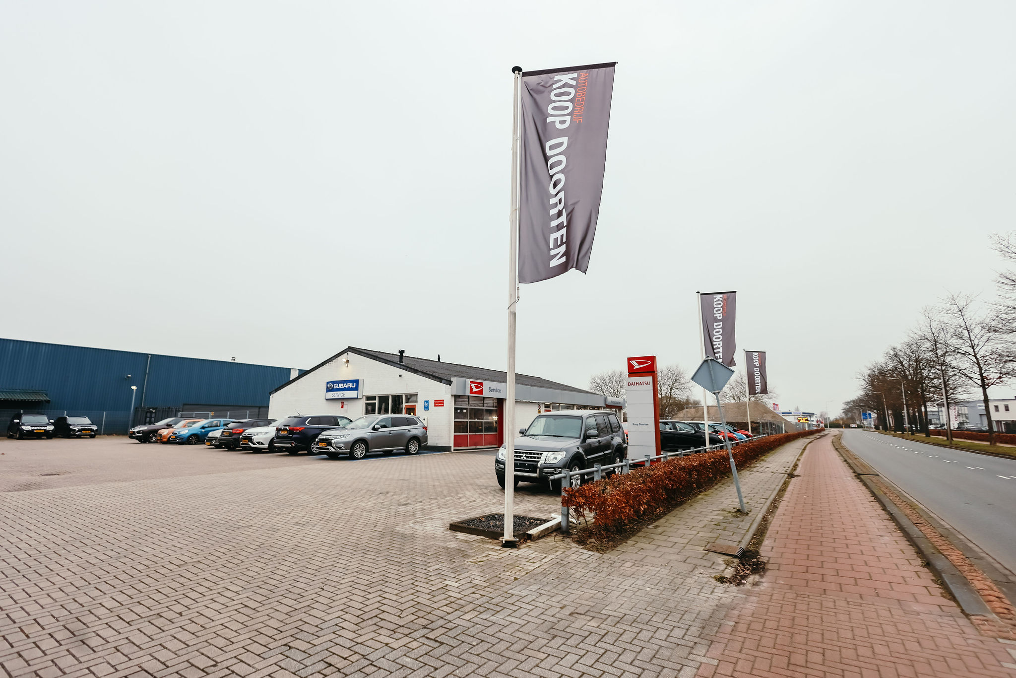 Het pand van Autobedrijf Doorten in Hoogeveen, met Subaru en Daihatsu serviceborden, een groot parkeerterrein vol auto's en vlaggen met "Koop Doorten" langs de weg. Op de voorgrond staat een zwarte SUV geparkeerd bij de ingang.