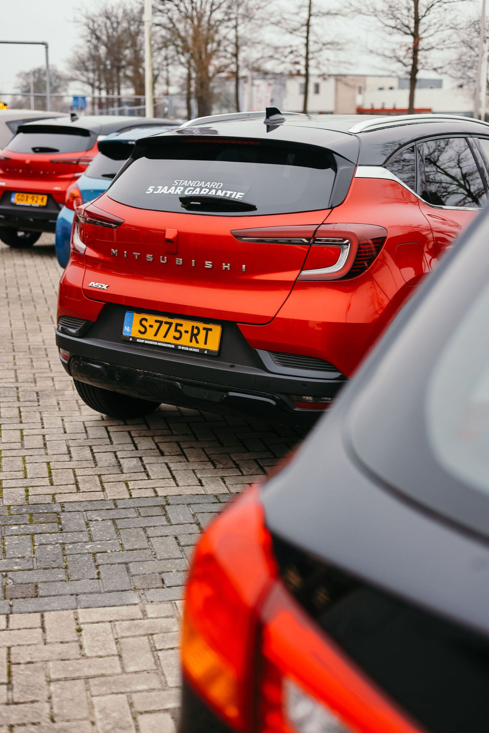 Een rode Mitsubishi ASX met een kentekenplaat "S-775-RT" staat op een parkeerplaats, met een sticker op de achterruit die "Standaard 5 jaar garantie" aangeeft, en andere auto's zichtbaar op de achtergrond.