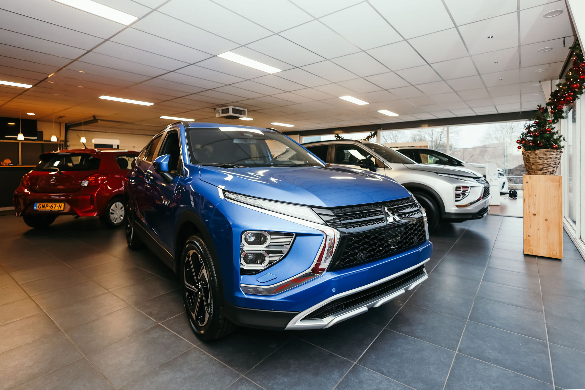 Een moderne showroom met drie auto's op display: een blauwe Mitsubishi SUV prominent op de voorgrond, een witte SUV ernaast en een rode hatchback op de achtergrond, met kerstdecoratie bij het raam.