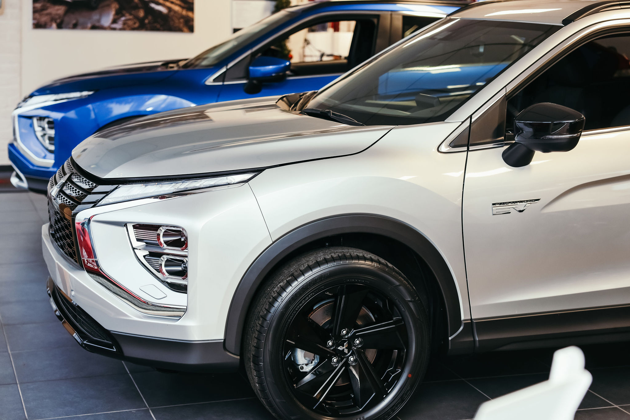 Close-up van de voorkant van een witte Mitsubishi SUV met een opvallend ontwerp, inclusief strakke koplampen en zwarte velgen, en een blauwe auto op de achtergrond in een showroom.