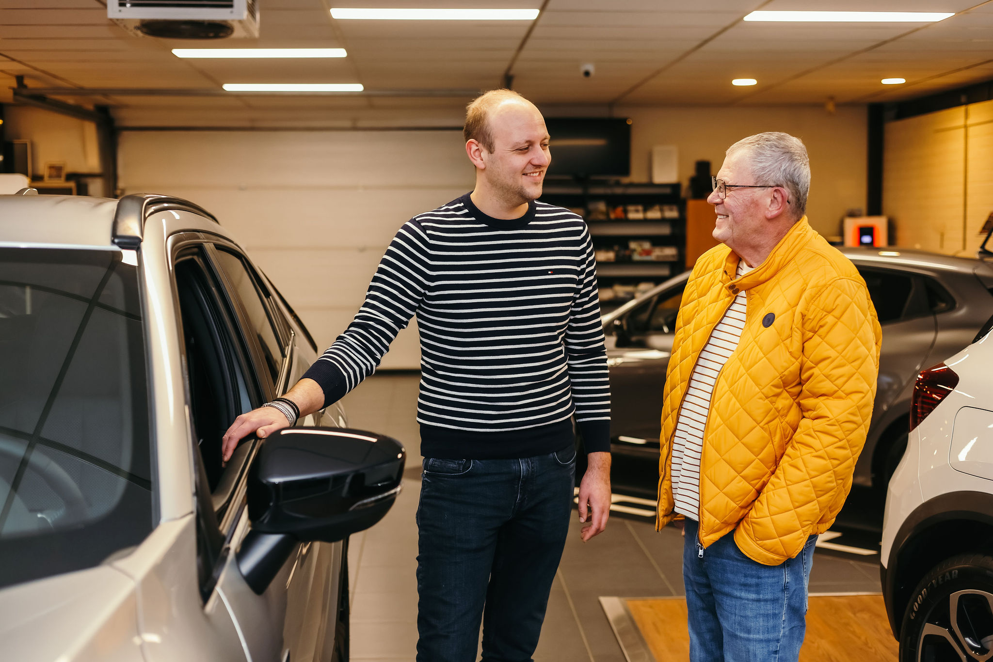 Een medewerker in een gestreepte trui en een klant in een gele jas staan lachend naast een auto in een showroom, in een ontspannen gesprek.