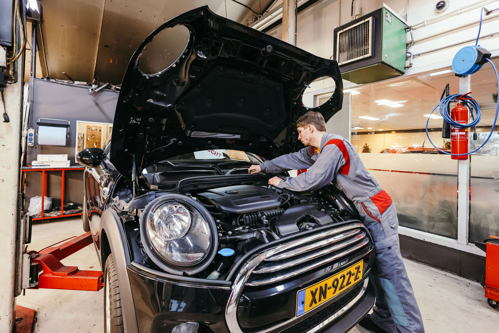 Een monteur in een grijze overall met rode accenten werkt onder de geopende motorkap van een zwarte Mini Cooper, die op een autobrug in een goed uitgeruste garage staat.