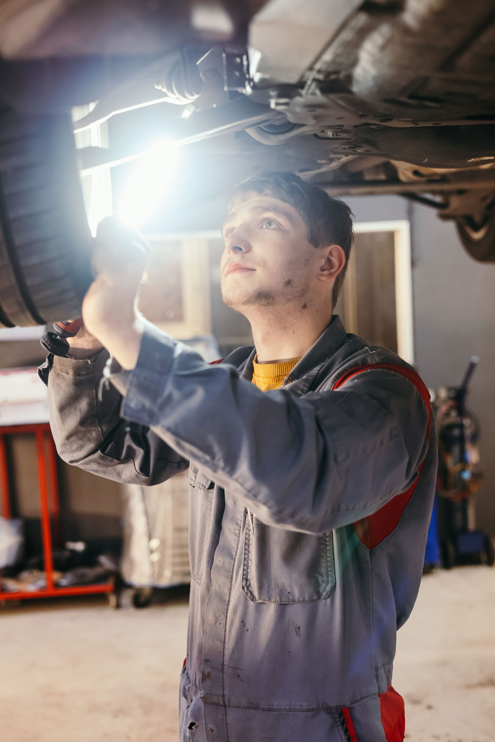 Een jonge monteur in een blauwe overall werkt aan de onderkant van een auto in een garage, met een felle lamp die het werkgebied verlicht.