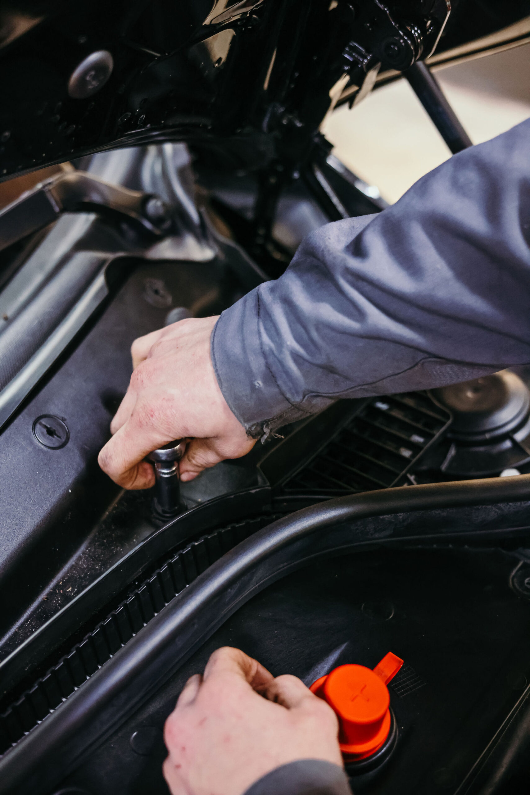 Close-up van de handen van een monteur die aan een auto werkt onder de motorkap, waarbij hij met gereedschap een onderdeel vastzet, met zichtbare kabels en een rood accudeksel.